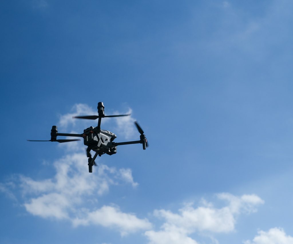 Picture showing drone flying in air against a blue sky with some clouds, taken from beneath the drone.