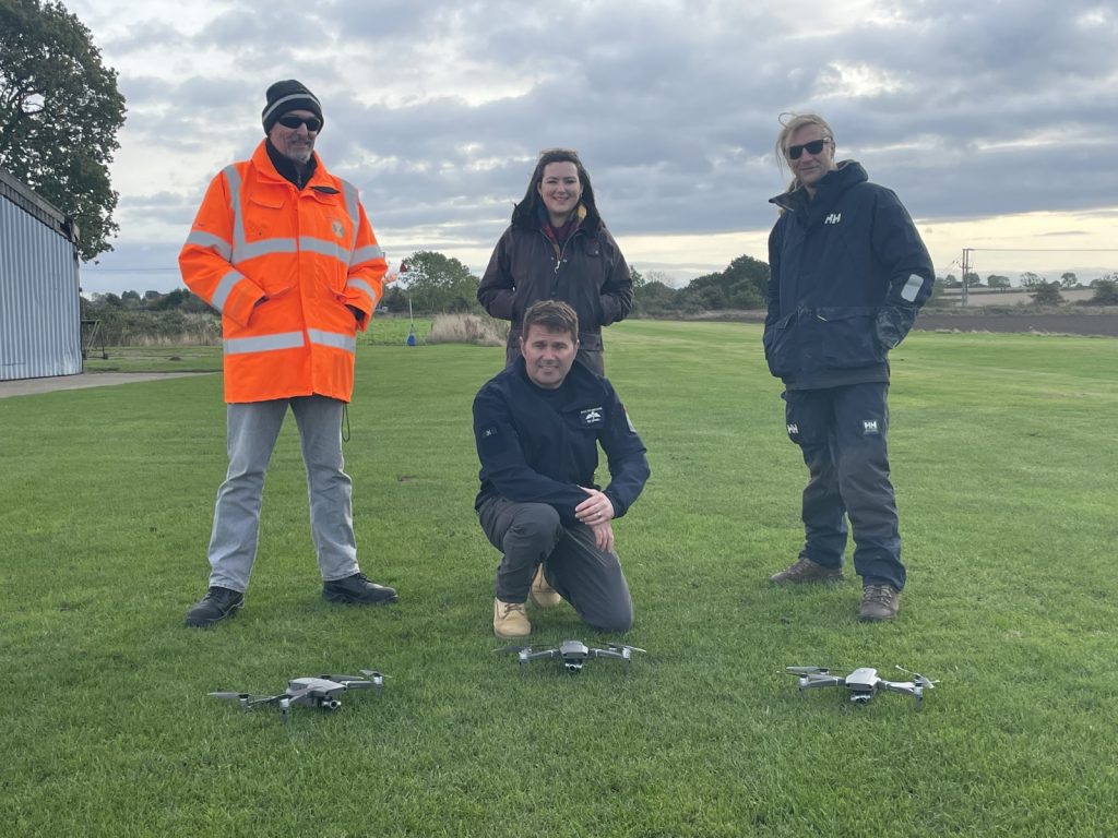 Man knelt down with three people standing and three drones on the grass in front.