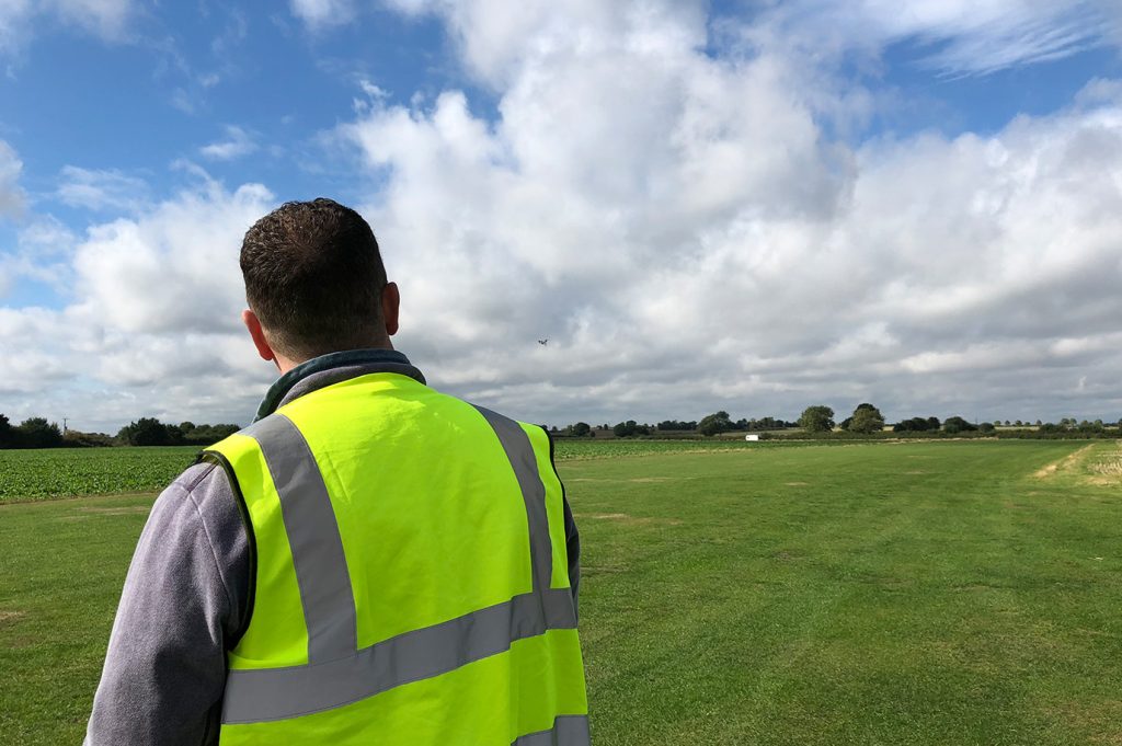 Image of man wearing high vis vest, shot from behind controlling a drone in the background.