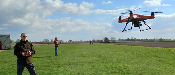 Man flying a drone in a field with the drone pictured at the top right of the image.