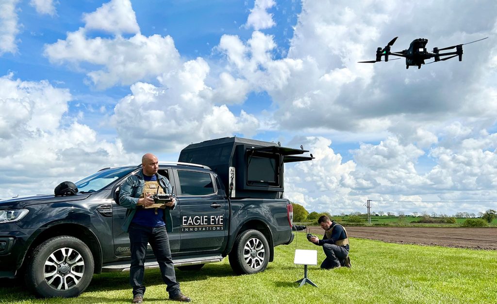 Man flying a drone in an armoured vest. Stood in front of a large truck with another man crouching down behind it.