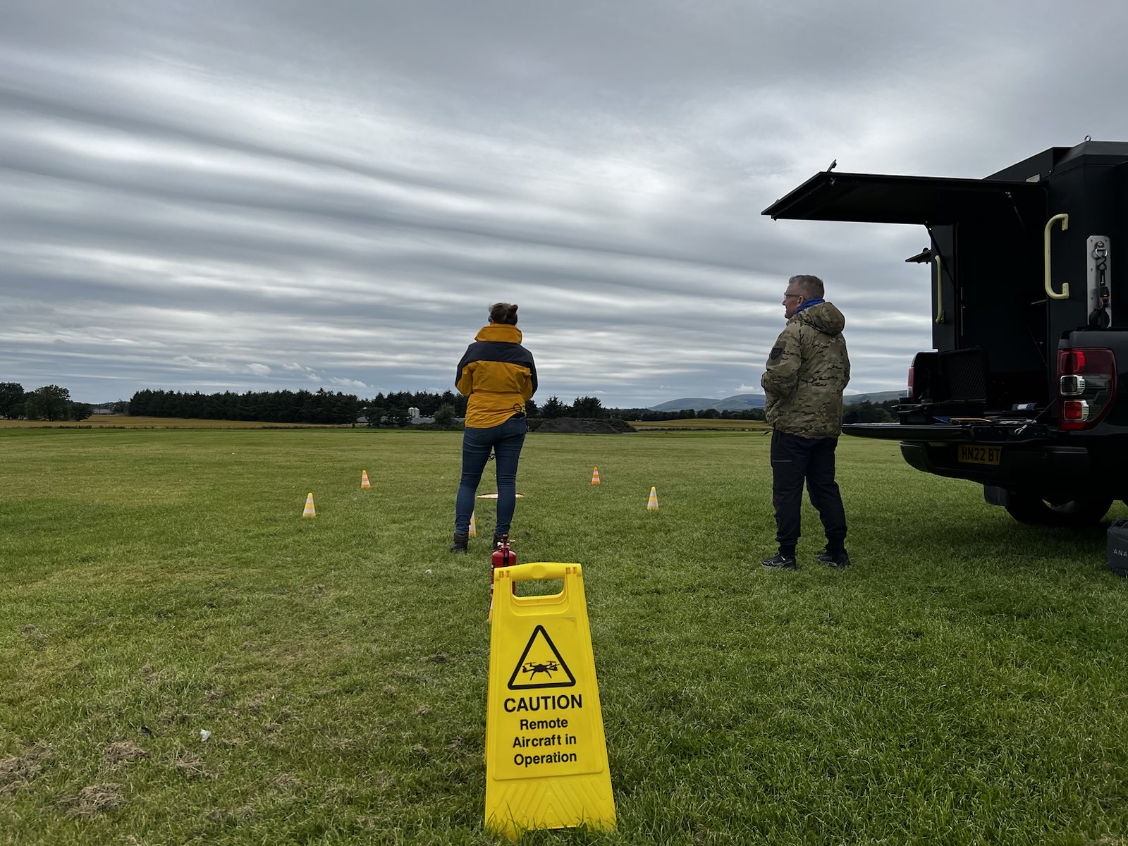 Two people standing in a field flying a drone, a warning sign outlines the importance of flight planning.