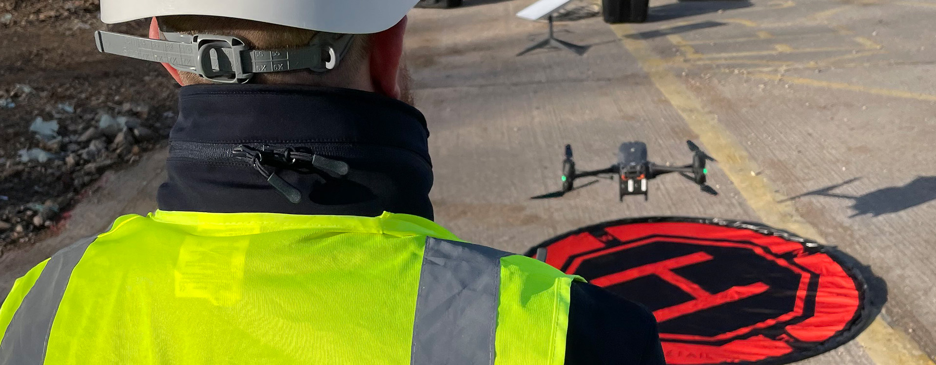 Man in high vis flying a drone above a landing pad at a construction site.