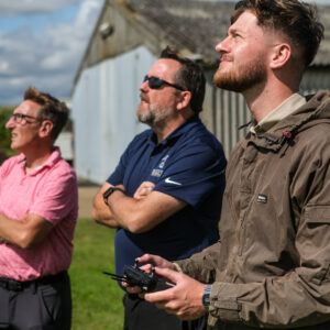 Three people stand outdoors watching a drone in flight. One person in the foreground holds a remote controller, while the others observe attentively beside him or an RPC course.