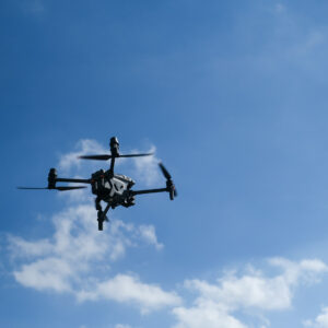 A professional quadcopter drone hovers mid-air against a clear blue sky with light clouds. The drone’s propellers are spinning, and its camera system is visible underneath, capturing imagery or data during flight.